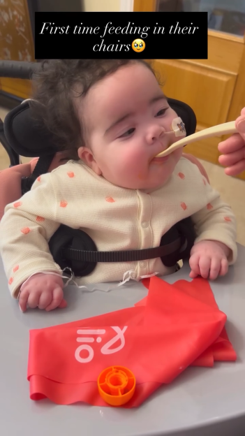 Baby in a feeding chair with a nasal tube and chest restraint, being fed with a spoon.