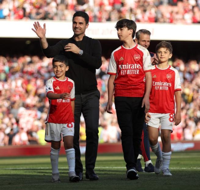 Mikel Arteta walks with three children in Arsenal uniforms on a soccer field.