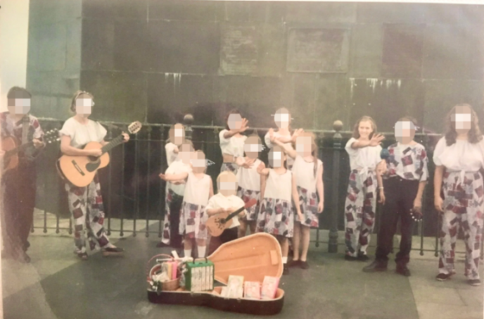Group of cult children singing at the base of the Cristo statue in Rio de Janeiro, with two adults playing guitars and a guitar case filled with offerings.
