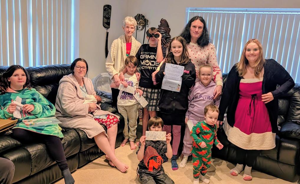 A family of 12 people gathered in a living room, smiling at the camera and holding up tickets and papers.