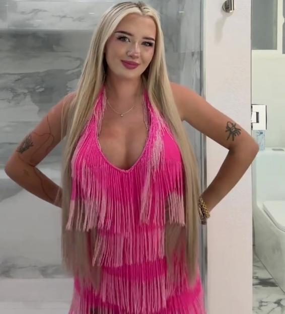 A young woman with long blonde hair and a pink fringed dress stands with her hands on her hips, smiling at the camera.