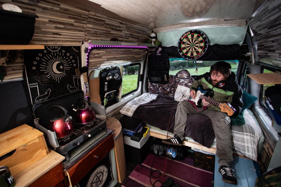 Tim Knight playing an electric guitar inside his converted minibus, which is also a home with a small kitchen and a bed.