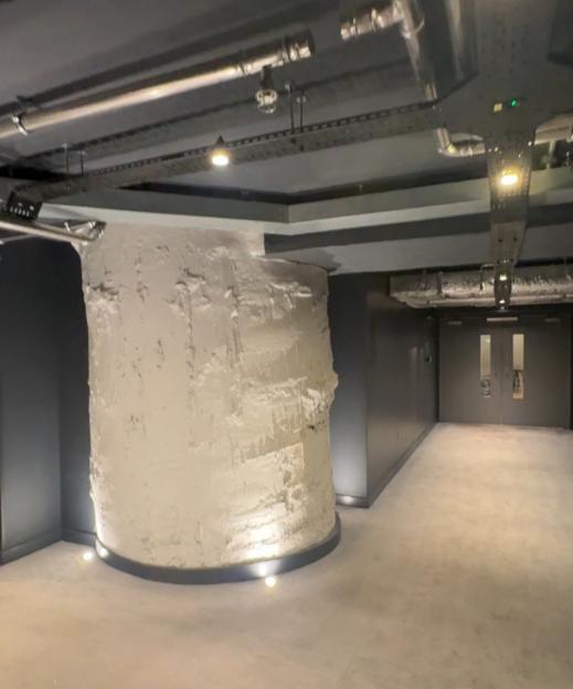 Interior view of a modern industrial space with a large white column, pipes on the ceiling, and a hallway leading to double doors.