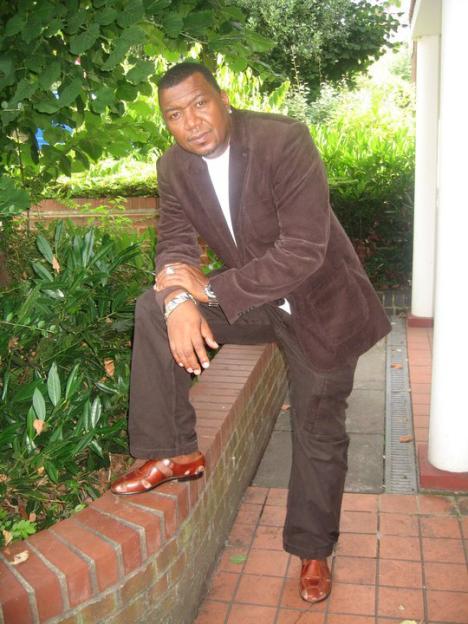 A man in a brown suit jacket, brown trousers, and brown leather shoes, posing with one foot on a brick wall.