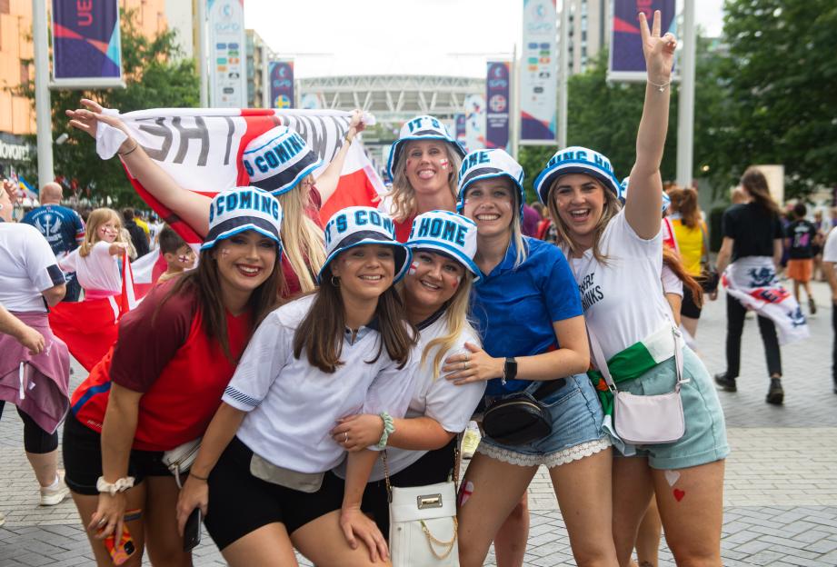 Six women wearing "It's Coming Home" hats and face paint, with England flags, cheering at the UEFA Women's EURO.