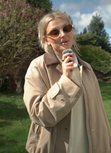 Woman in sunglasses and a beige jacket speaking into a microphone outdoors.