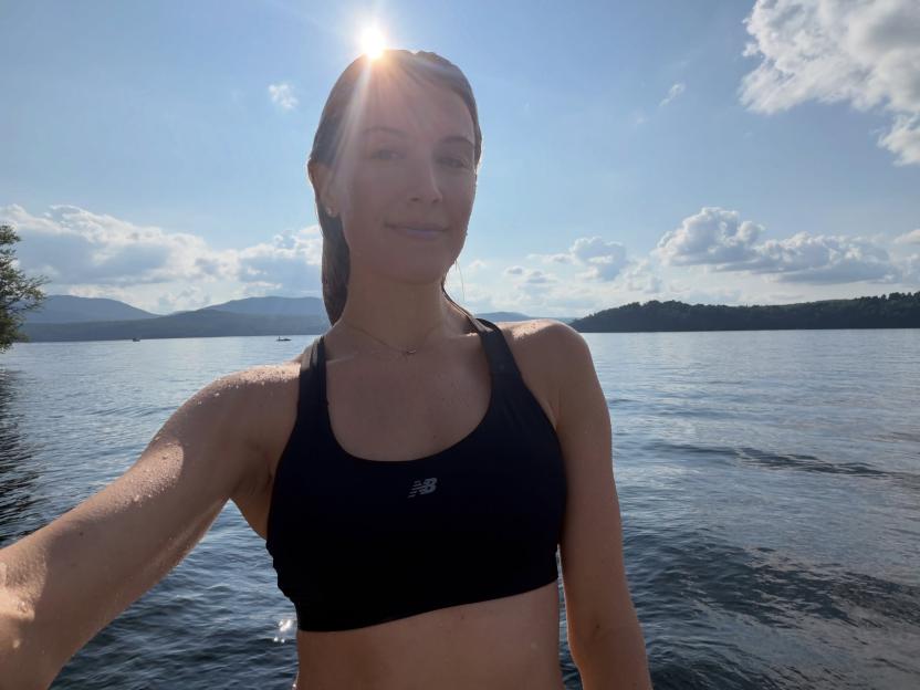 A woman in a black sports bra, wet from swimming, standing in a lake with mountains and forests in the background.