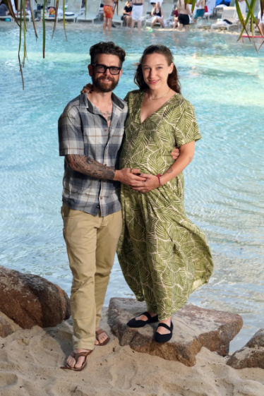 Man with arm around a smiling pregnant woman in a green dress, standing on a sandy shore by blue water.