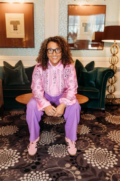 A woman with curly hair, wearing a pink bandana-print shirt and purple pants, smiles while sitting on a coffee table.