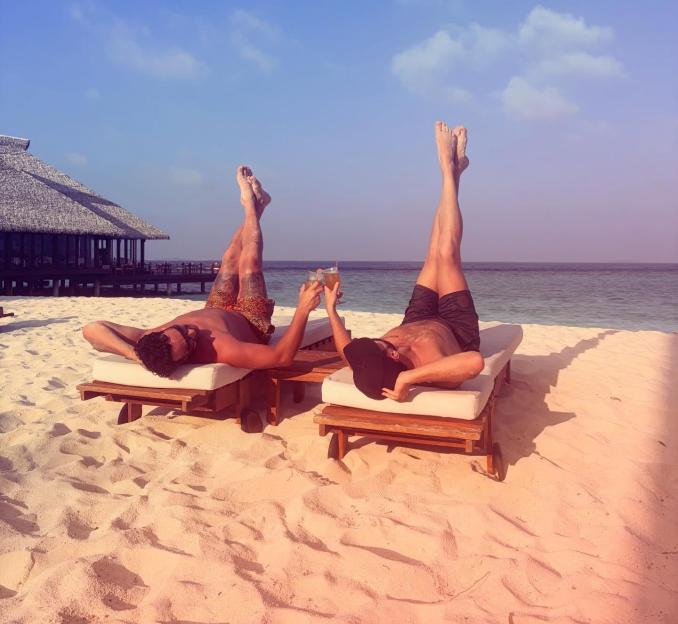 Two men relaxing on lounge chairs on a sandy beach with their legs raised, holding drinks, with the ocean and a building in the background.