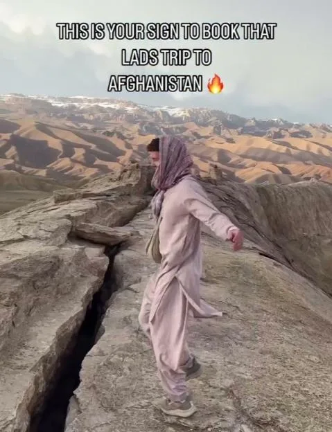 A man wearing traditional Afghan clothing walks along a rocky mountain ridge in Afghanistan, with text overlaying the top that reads "THIS IS YOUR SIGN TO BOOK THAT LADS TRIP TO AFGHANISTAN."