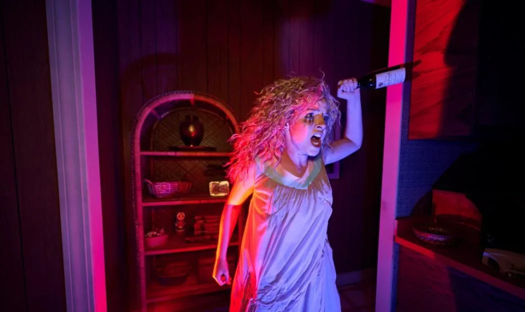 A woman with wild, curly hair and a distressed expression holds a wine bottle aloft in a dimly lit room with red and blue lighting.