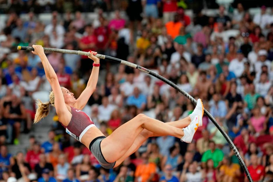 A female pole vaulter wearing a maroon and white top and dark shorts in mid-air holding onto the pole.