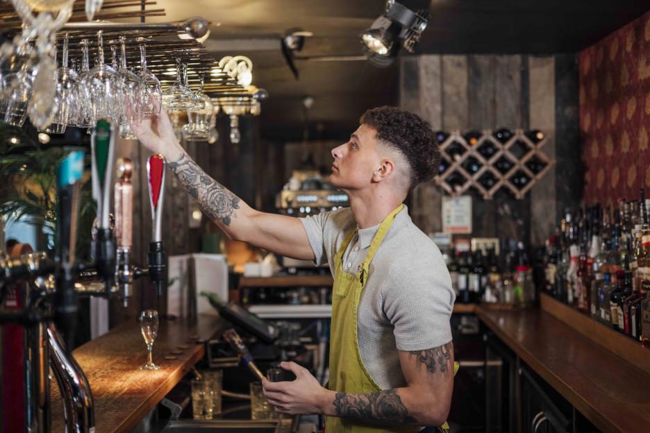 Bartender in an apron reaching for a wine glass above the bar.