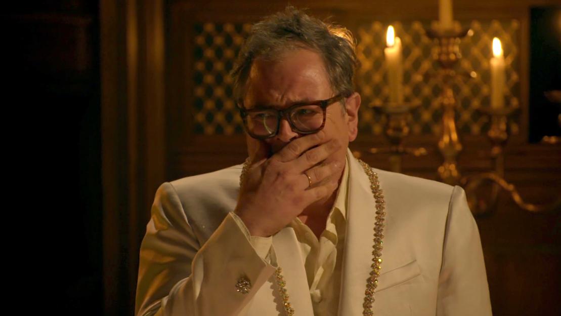 Dan Aykroyd as a rich man in a white suit covering his mouth with his hand.