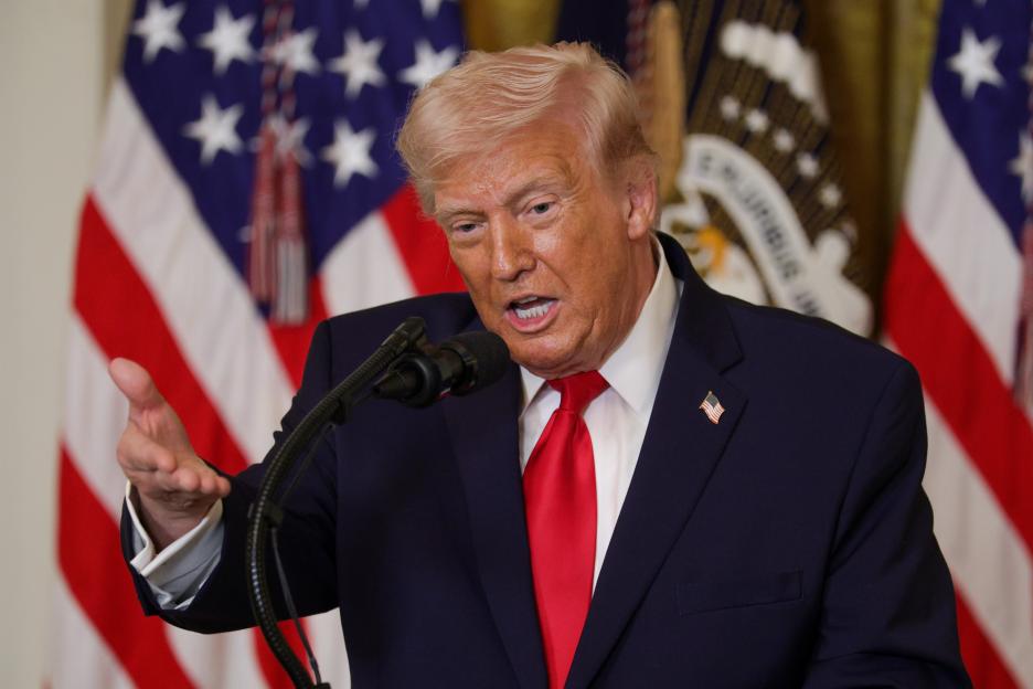 Donald Trump speaking at a Women's History Month event, with American flags and the presidential seal in the background.