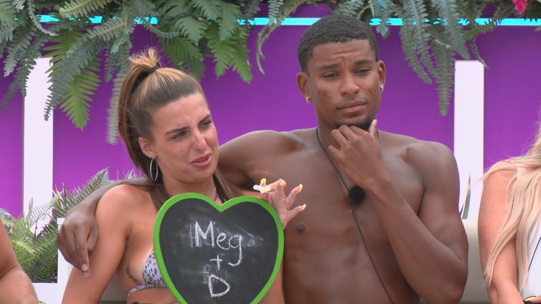 A couple, Toni and Cacha, on the television show "Love Island," with Toni holding a heart-shaped chalkboard that says "Meg + D."