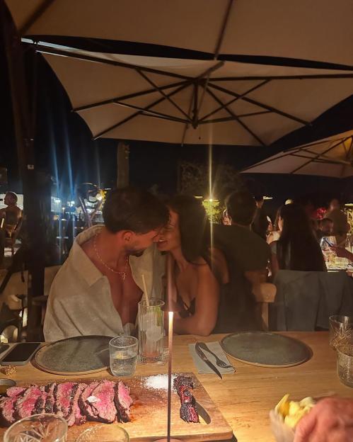 A couple leans in for a kiss at a restaurant table, with sliced steak and drinks in front of them.
