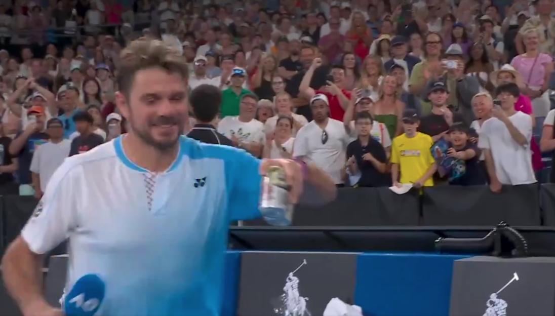 Tennis player Stan Wawrinka smiling and holding a drink on the court after a match, with cheering fans in the stands behind him.
