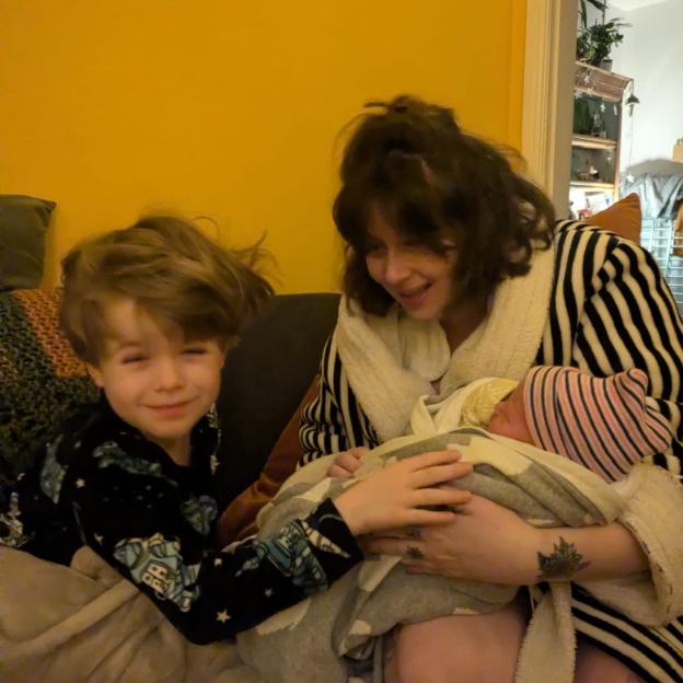 A woman in a striped robe holding a newborn and a young boy smiling beside her.
