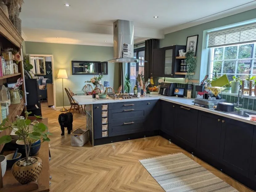 A large open kitchen with dark blue cabinets, sage green walls, a kitchen island, and light wood herringbone flooring. A black dog stands on the floor next to the island.