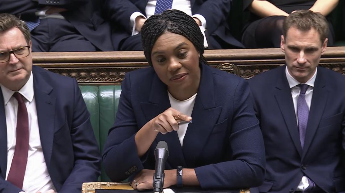 Kemi Badenoch speaks at a microphone in Parliament, with two men seated beside her.
