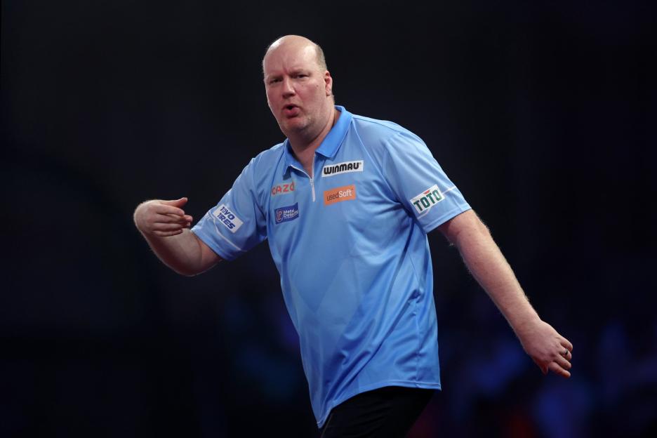 A male darts player in a light blue polo shirt with sponsor logos, gesturing with his left hand, at the 2022/23 Cazoo World Darts Championship.