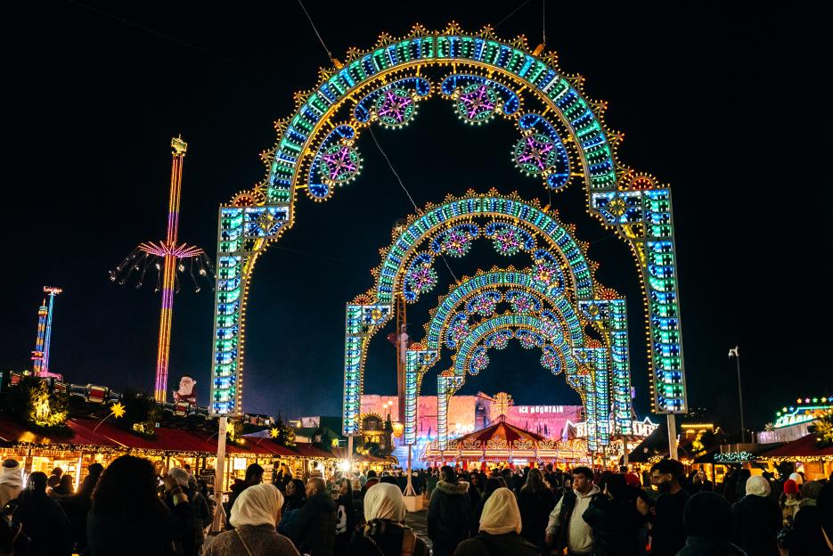 Crowds of people at Winter Wonderland at night, with multiple ornate arches adorned with multicolored lights spanning over them, and several amusement park rides visible in the background.