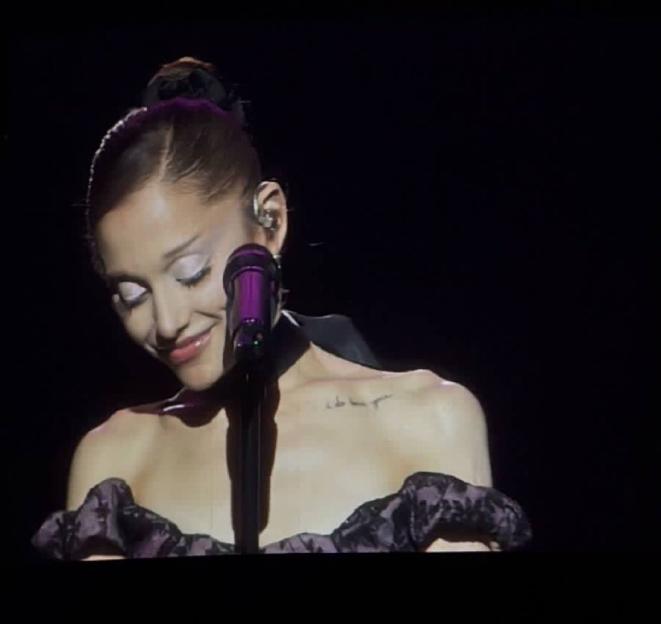 Ariana Grande smiles as she performs, with a tattoo visible near her left collar bone.