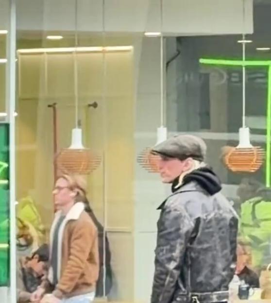 Two men walking past a storefront, one in a brown jacket and the other in a dark jacket and flat cap.