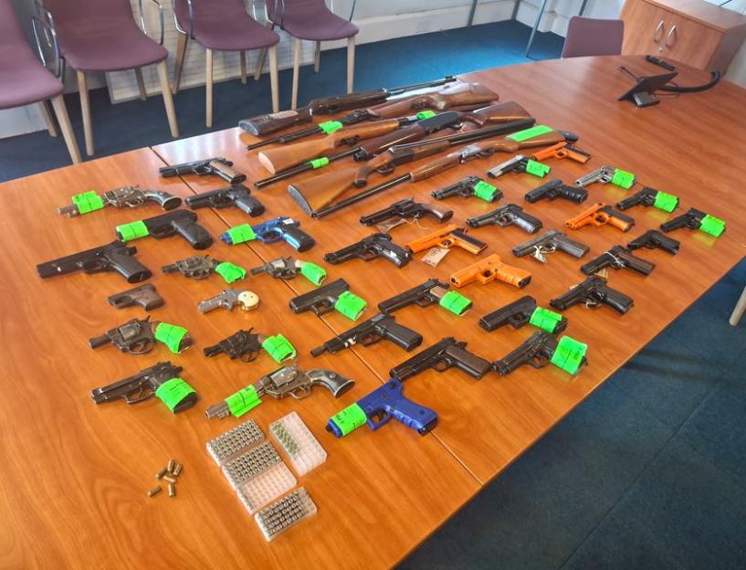 Various firearms, including pistols, revolvers, and rifles, displayed on a wooden table, some with green tags and ammunition.
