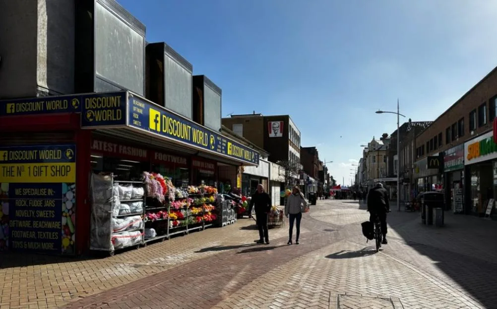 A pedestrianised high street in Rhyl with shops on either side, including a 'Discount World' storefront, with a few people walking and cycling on a sunny day.
