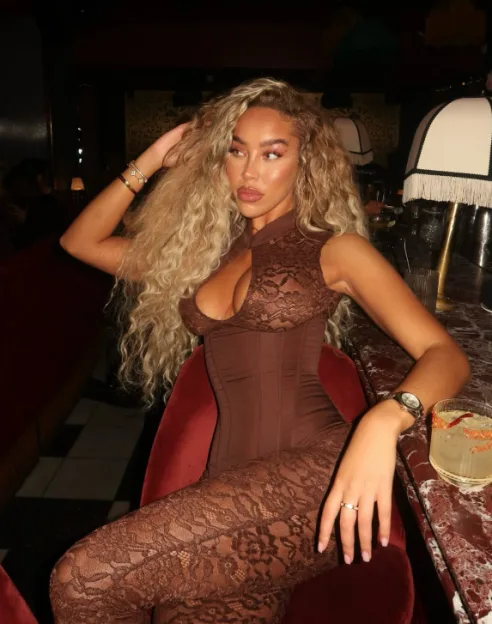 A woman in a brown lace jumpsuit with long blonde curly hair sitting on a red velvet chair.