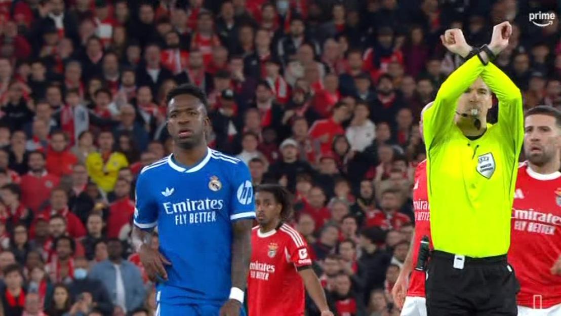 Football referee signaling "stop play" with crossed arms, while players and crowd look on.