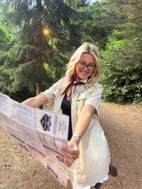 A smiling woman wearing a scout uniform and glasses, holding open a "Camp Wildfire Camper's Guide" that displays event schedules and "10th Anniversary" text.
