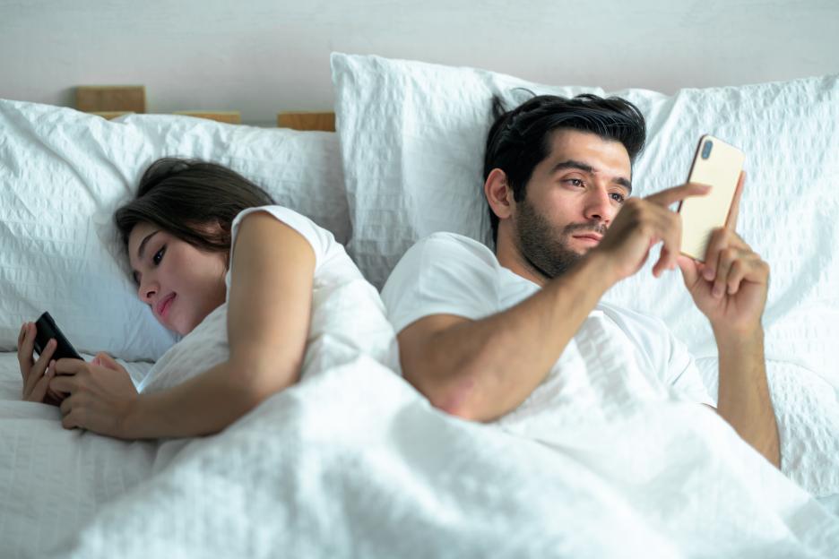 Couple in bed, backs turned to each other, individually using cell phones.