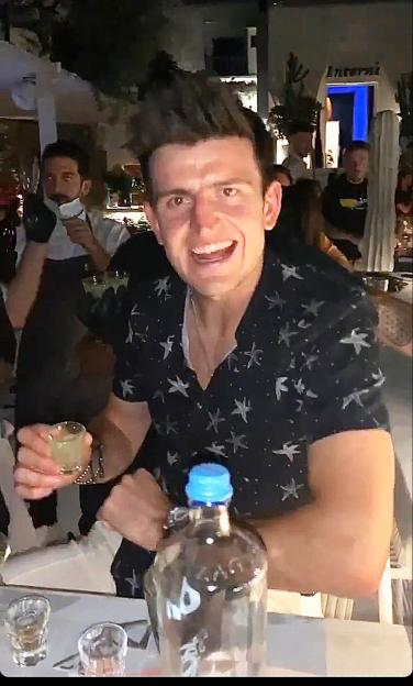Harry Maguire in Greece holding a shot glass, with an open bottle of liquor in front of him.