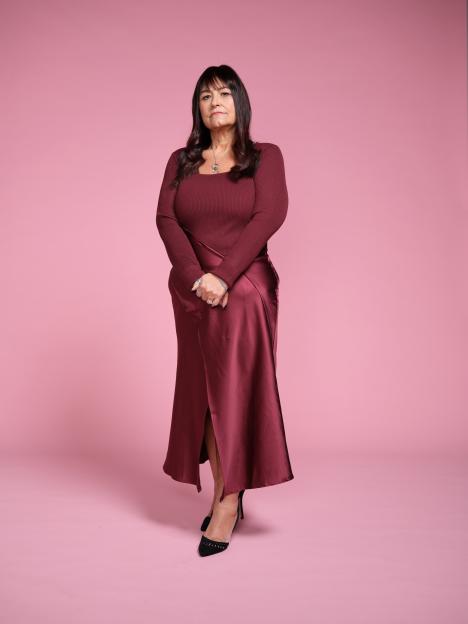 A woman with dark hair wearing a maroon long-sleeved top and a matching long skirt with a slit, standing against a pink background.