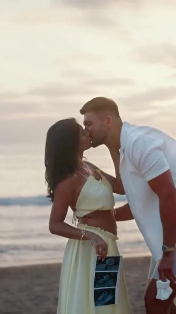 A couple kissing on a beach, the woman holding ultrasound photos and the man holding baby booties.