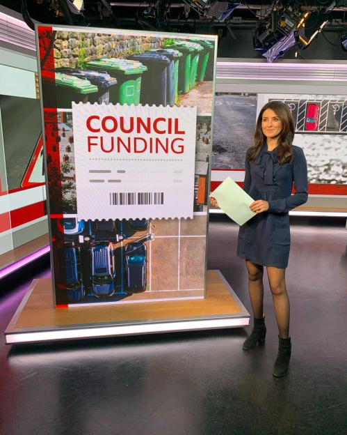 News presenter in a dark dress standing next to a screen displaying "Council Funding" with images of green bins and parked cars.
