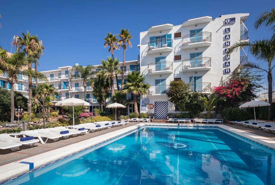 An outdoor swimming pool surrounded by lounge chairs, palm trees, and a white hotel building named "LANAMAR".