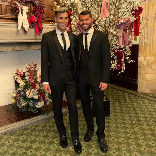 Junaid Ahmed and his fiancé, dressed in black suits and ties, stand smiling in front of a decorated fireplace and a blossoming tree.