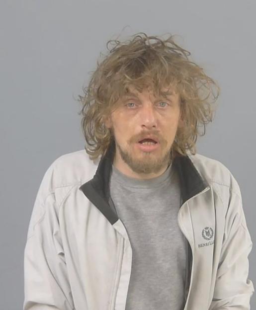 Mugshot of a man with disheveled brown hair and a beard, wearing a light-colored jacket over a gray t-shirt.