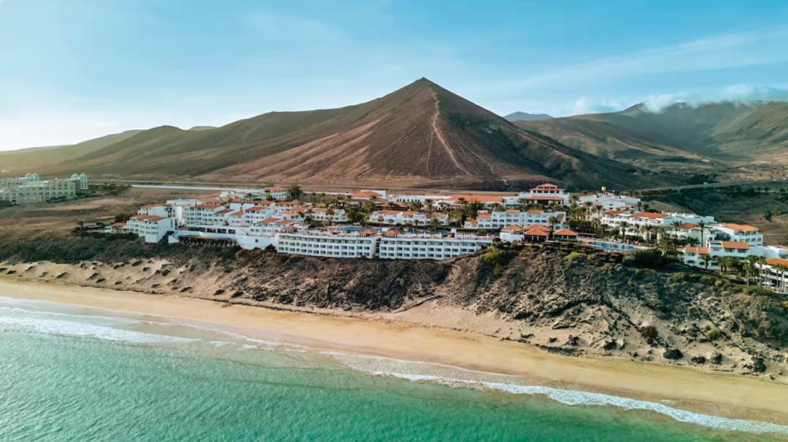 Aerial view of a coastal resort with white buildings and red roofs, backed by mountains.