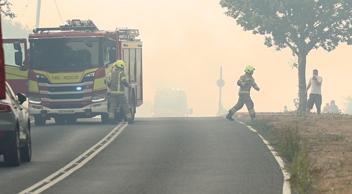 Firefighters and fire truck surrounded by smoke on a road, with onlookers in the background.