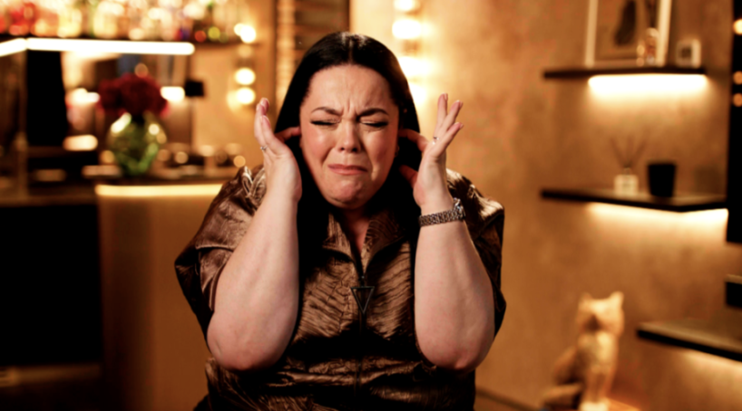 A woman with dark hair covering her ears with her hands, eyes closed and face scrunched in fear or disgust.