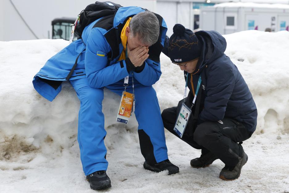Mykhailo Heraskevych, father of disqualified Ukrainian skeleton racer Vladyslav Heraskevych, covers his face in distress, with another person crouching beside him, on a snowy ground.