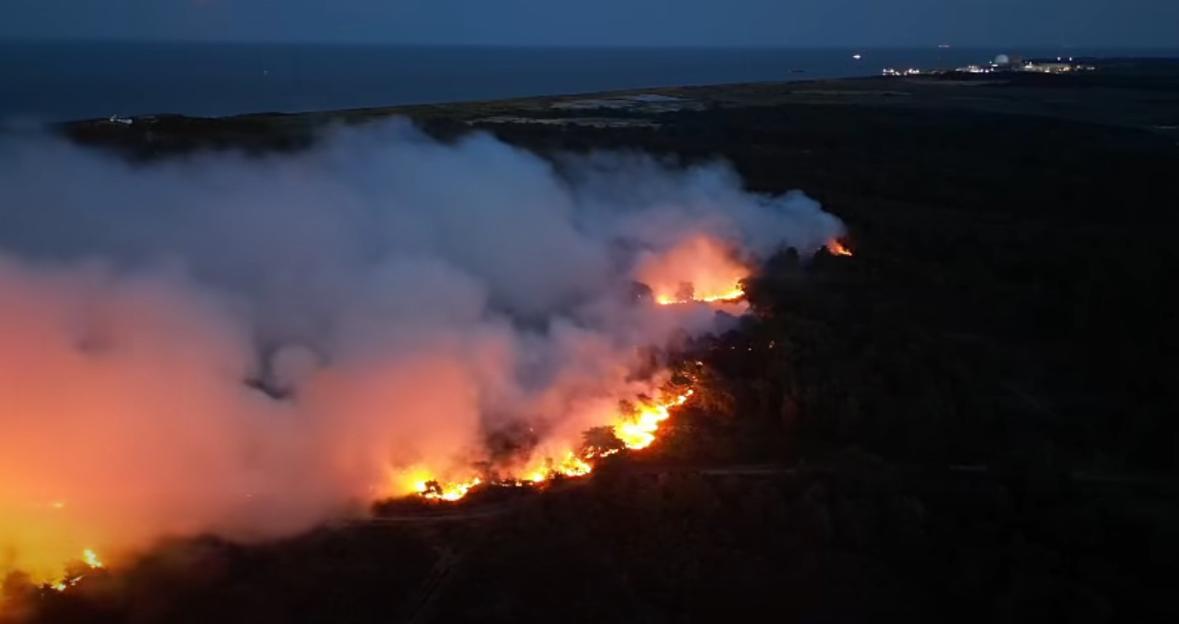 An aerial view of a nighttime wildfire with flames consuming trees and thick smoke rising, near a body of water and an industrial facility.