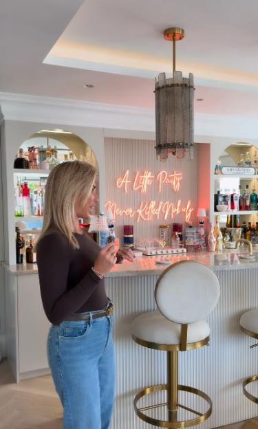 Billie Faiers stands in her garden cocktail bar with a "A Little Party Never Killed Nobody" neon sign.