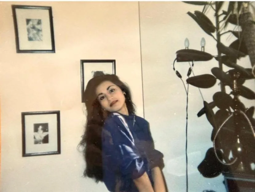 A vintage photo of Jinda Mann in her youth, posing indoors with framed pictures on the wall and a large houseplant beside her.